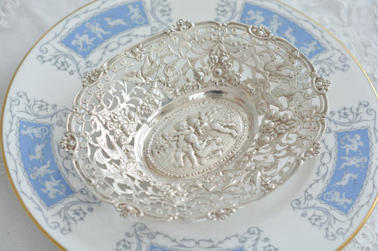 天使にリボンと薔薇のガーランドのピアス細工がとても愛らしく美しいボンボンディッシュです。 こちらはドイツで作られてイギリスに輸入されたお品です。繊細なピアス細工(透かし模様)に可愛らしい天使達がとても美しいお品です。ボンボンディッシュとはチョコレートやキャンディーなど小さなお菓子を入れて、ゲストがつまめるようにテーブルなどに置いておくためのお皿で、テーブルやお部屋に置かれていました。小ぶりなお皿で、ちょっとしたお菓子を乗せるのにぴったりです。