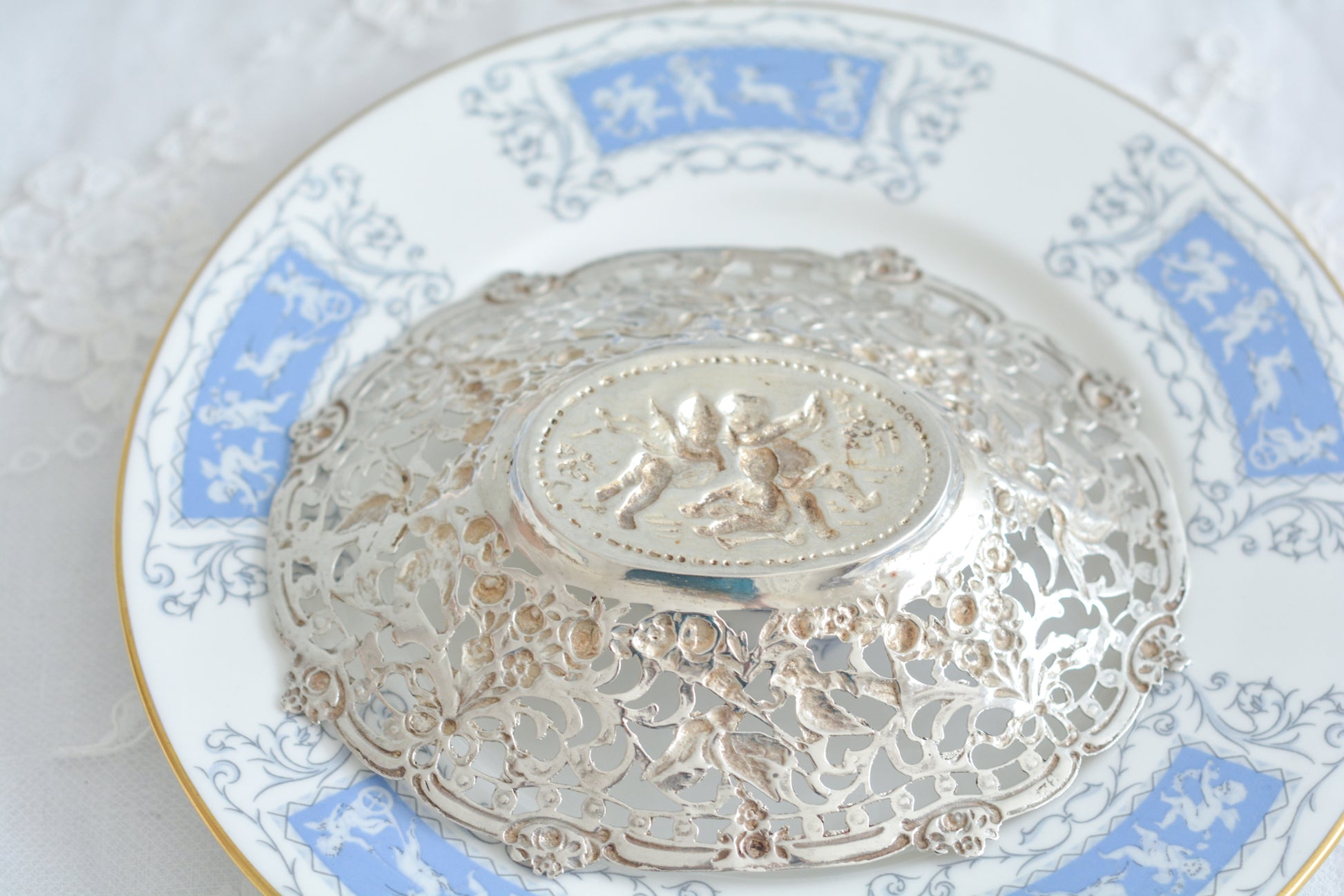 天使にリボンと薔薇のガーランドのピアス細工がとても愛らしく美しいボンボンディッシュです。 こちらはドイツで作られてイギリスに輸入されたお品です。繊細なピアス細工(透かし模様)に可愛らしい天使達がとても美しいお品です。ボンボンディッシュとはチョコレートやキャンディーなど小さなお菓子を入れて、ゲストがつまめるようにテーブルなどに置いておくためのお皿で、テーブルやお部屋に置かれていました。小ぶりなお皿で、ちょっとしたお菓子を乗せるのにぴったりです。